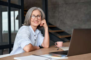 Smiling mature middle aged woman sitting at workplace with laptop, portrait.